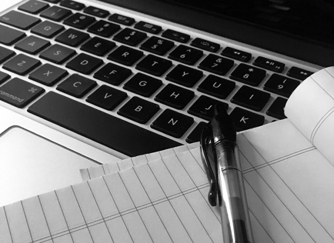 Black and White Computer Keyboard Grayscale image of a computer keyboard with black keys. Visible atop the keyboard is a portion of a notepad and the top of a black and clear barrelled ink pen.